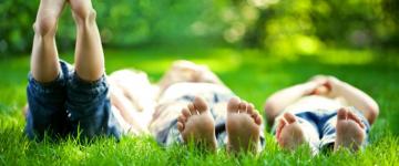 Photo of three kids lying barefoot in the grass. Their feet are close to the camera. One kid has his feet raised to the sky.