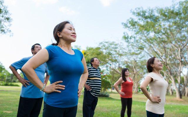 Photo of a group of adults outdoors, each striking the same yoga pose with their hands on their hips and eyes closed.