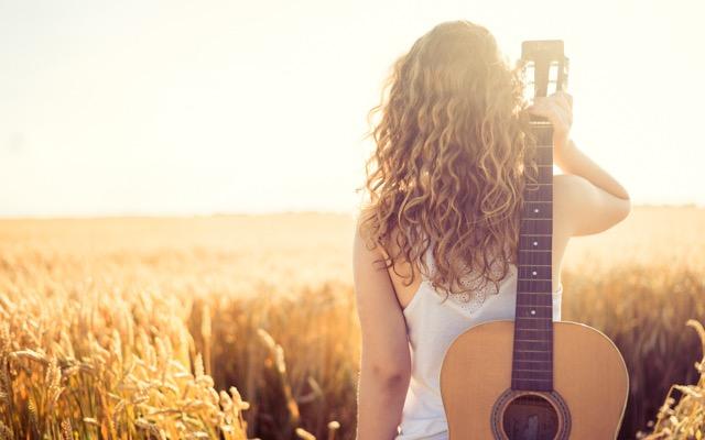 Photo of a woman facing away from the camera, looking out over a field as she holds an acoustic guitar over her shoulder.