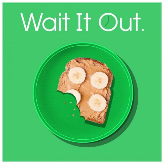 Open-face peanut butter and banana sandwich with a few bites taken out of it on a plate. Text above it says "Wait it Out." The O of the word "out" has the hands of a clock inside it.